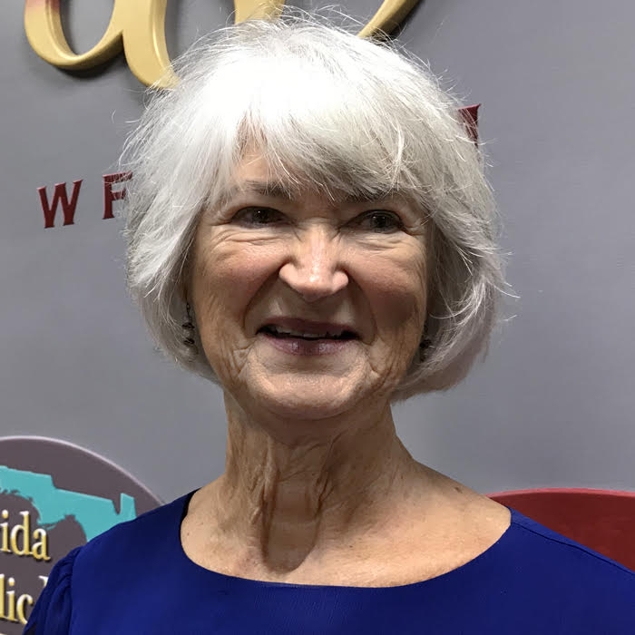 A smiling older, white haired woman in a blue blouse in front of a light purple wall.