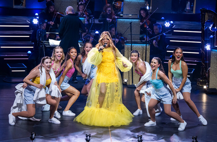 woman in yellow singing, surrounded by dancers
