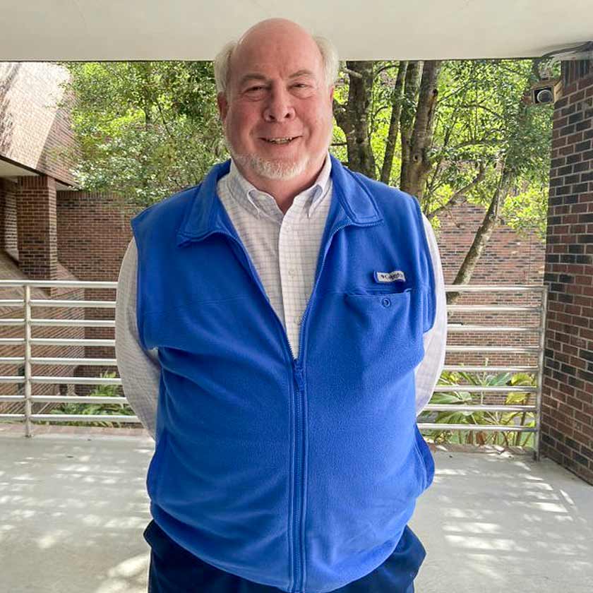 A smiling man in a blue vest stands outside.