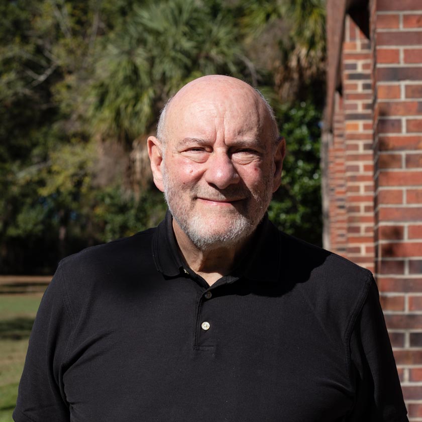 A man in a black shirt stands outside in front of greenery and a red-brick wall.