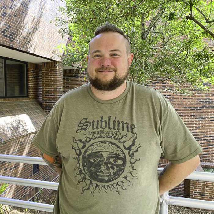 A smiling man stands in front of shrubbery.