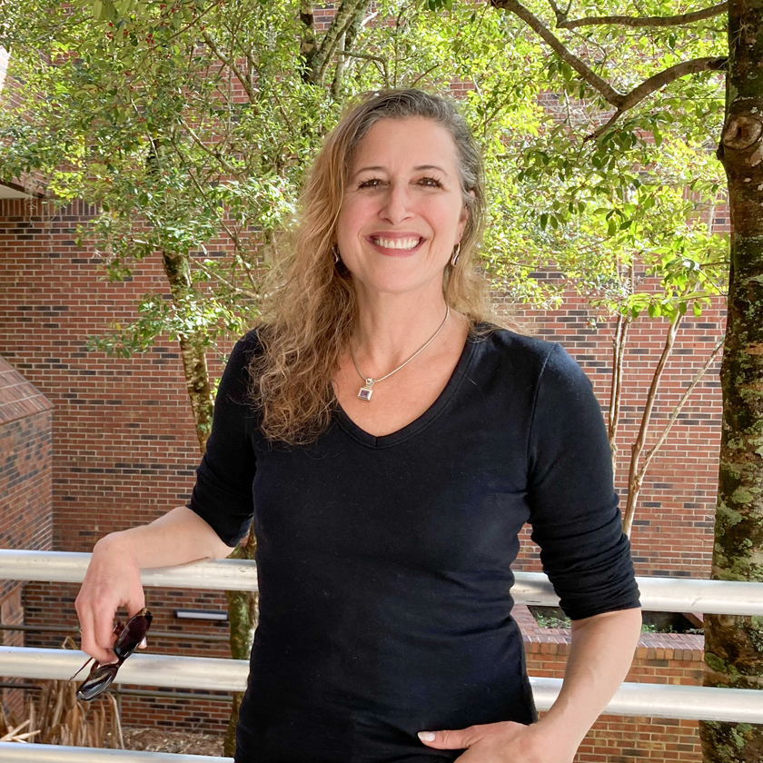 A smiling woman stands in front of a red-brick wall and a tree.