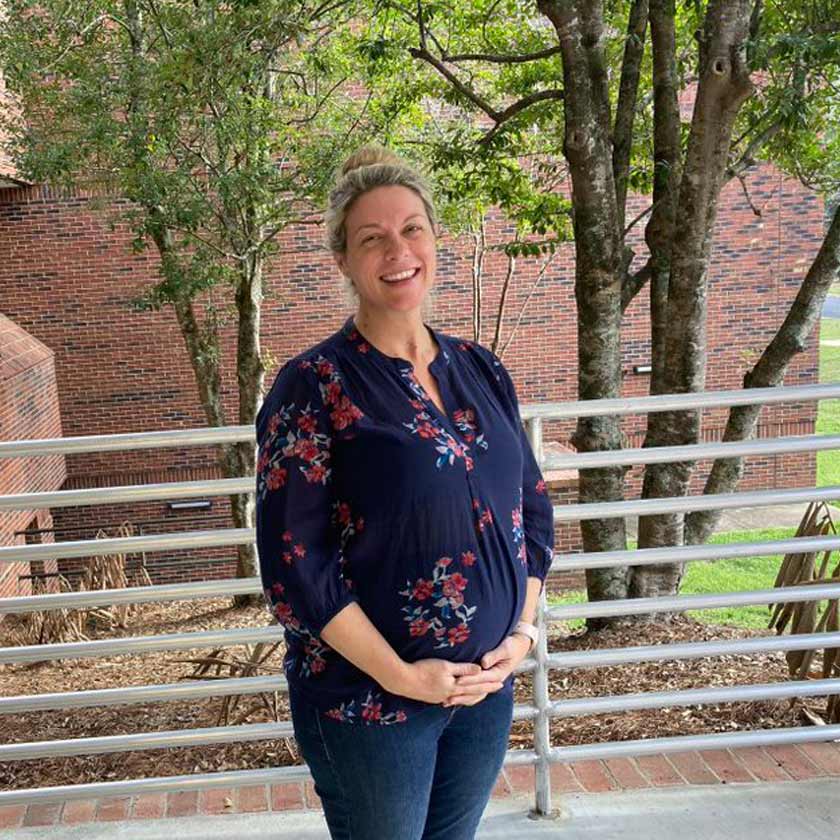 A smiling woman stands in front of shrubbery.